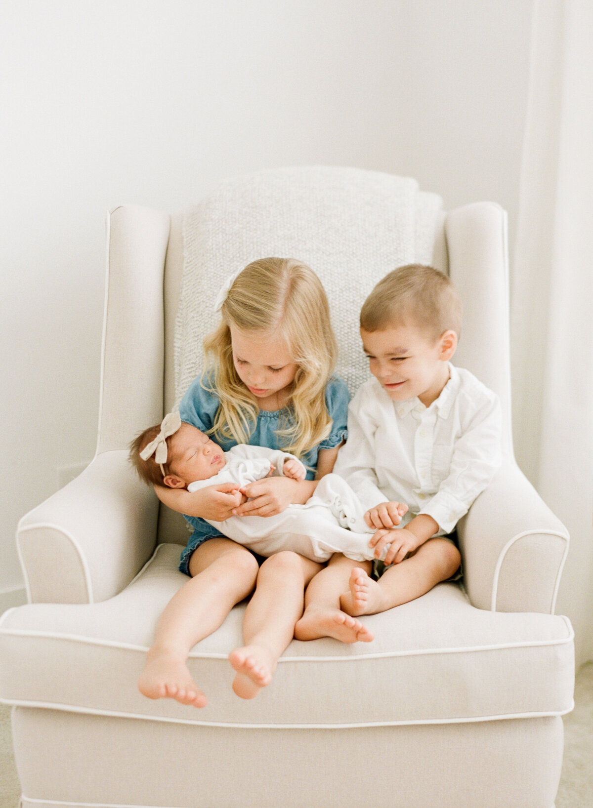 Big siblings sit in a white rocker while holding their newborn sister during a newborn session photographed in Raleigh. Photographed by newborn photographer Raleigh A.J. Dunlap Photography.