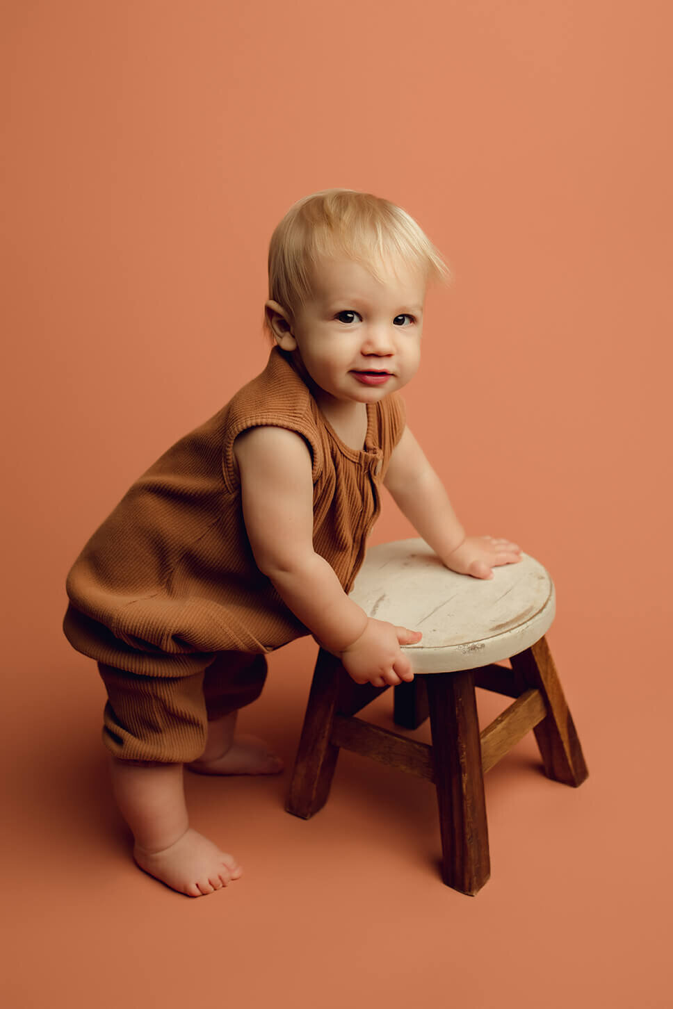 a one year old boy standing on a brown drop wearing a brown outfit holding a brown and white stool