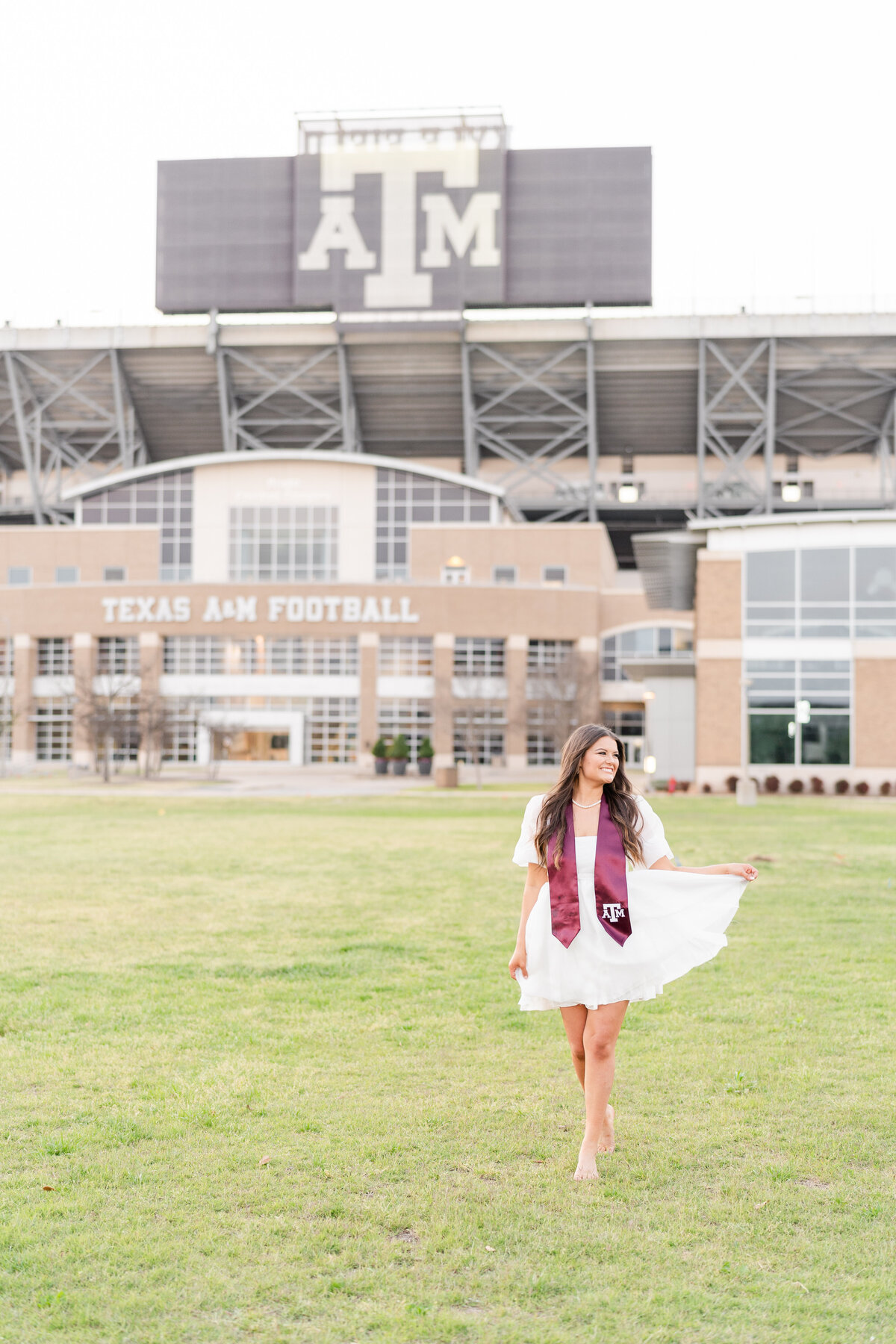 Texas A&M senior girl running through field in fluffy white dress while wearing Aggie stole with Kyle Field in background