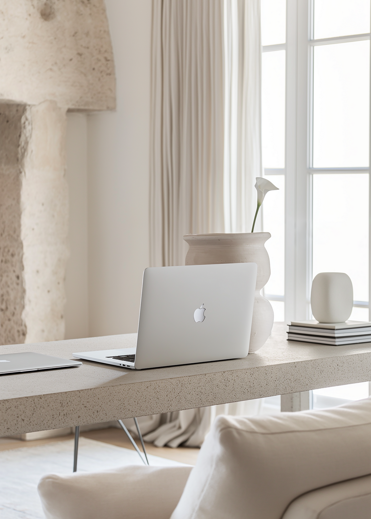 A bright and airy interior design photograph showcasing a contemporary minimalist office setup for a fashion editor. A MacBook sits on a sleek desk, surrounded by stone accents and neutral colours. A stack of design magazines titled "Iconic Works of Design" adds to the sophisticated aesthetic.