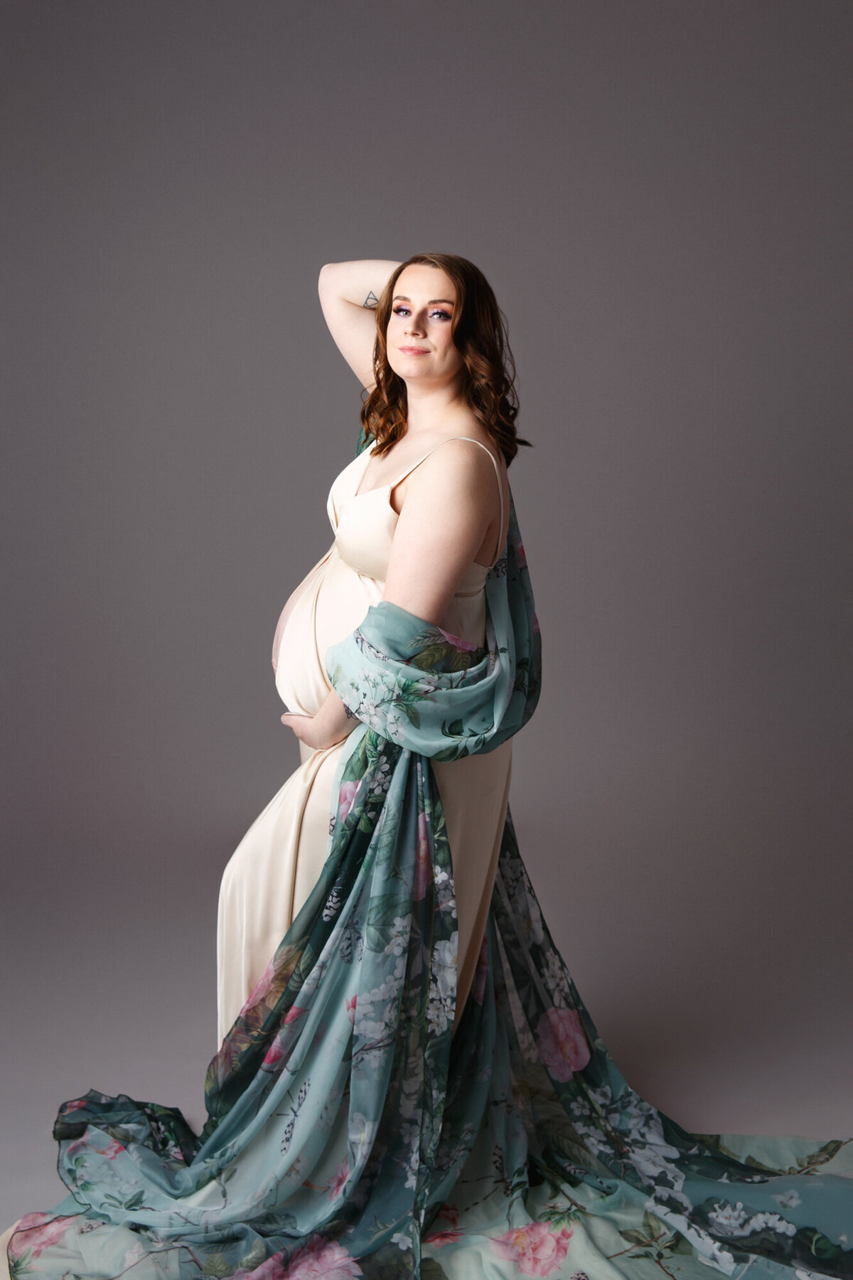 Full body portrait of a w pregnant woman wearing a gold dress with a green floral scarf wraped in her arms and photographed on a dark gray background