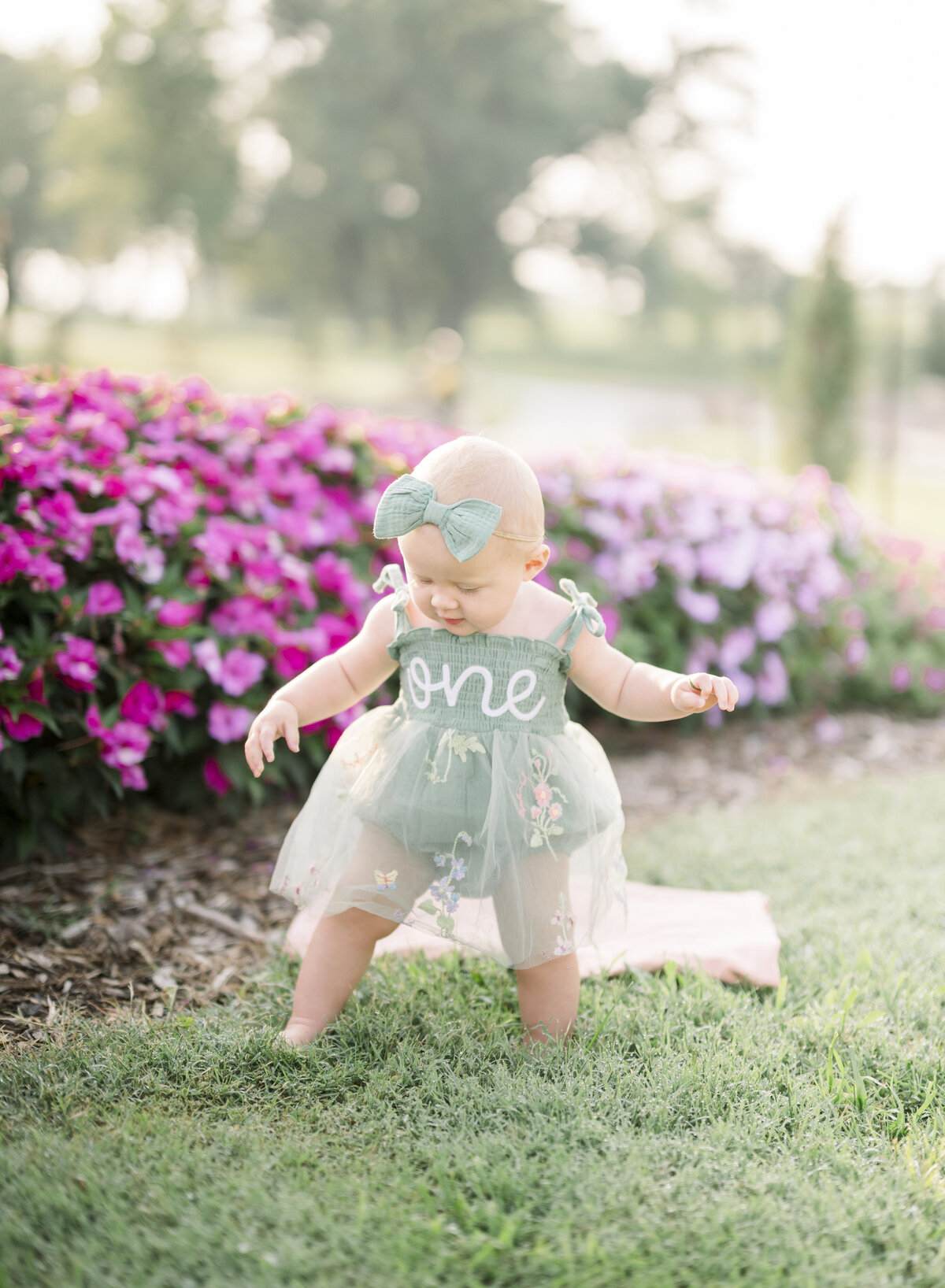 Baby girl standing in the grass near vibrant pink flowers during an outdoor Kansas City milestone session.