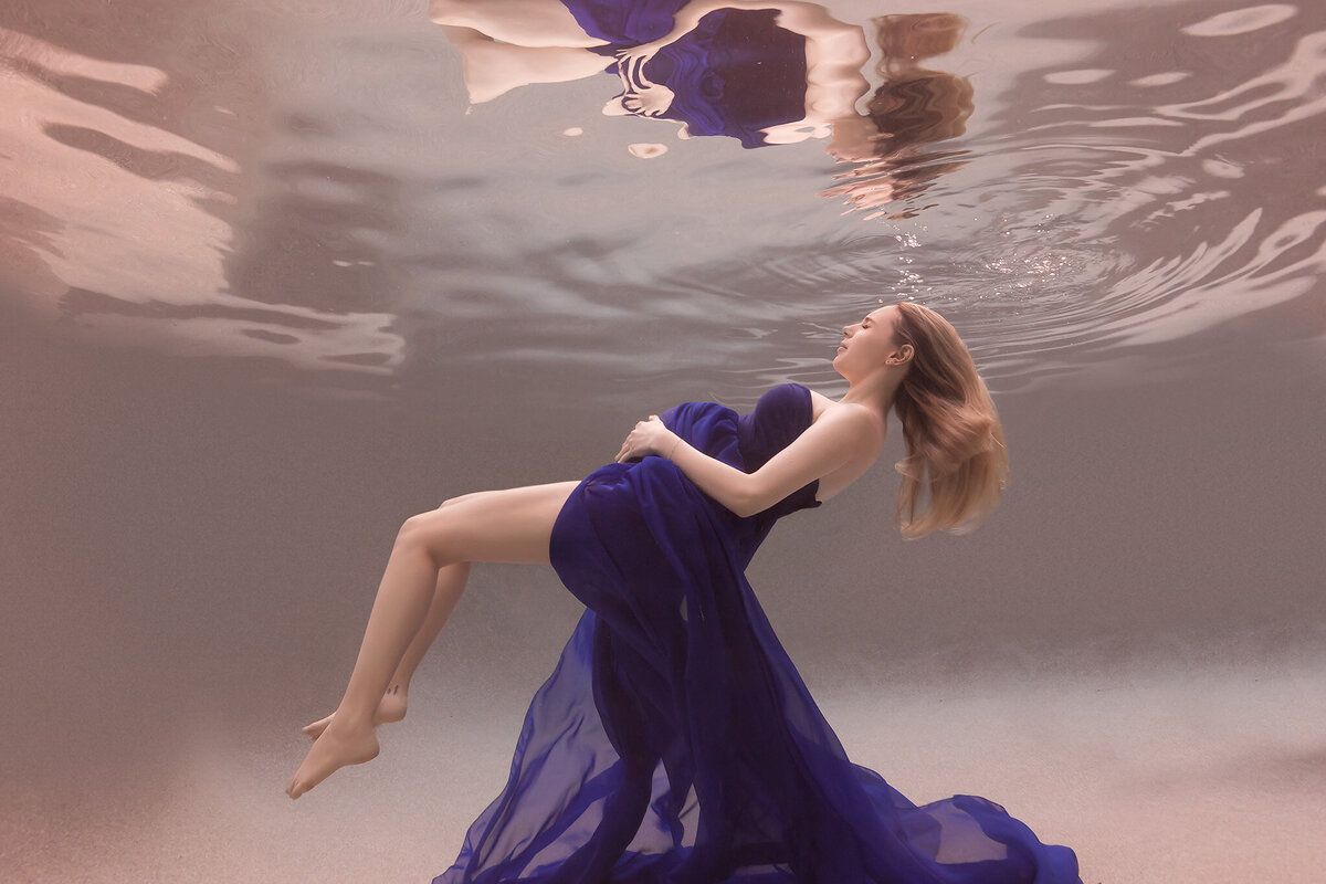 A serene underwater maternity photo captured by a Dallas underwater maternity photographer. The woman floats effortlessly in the water, her rich blue gown cascading around her like waves. Her hair fans out gracefully as she cradles her belly with a calm, blissful expression, showcasing the beauty of underwater maternity photography.