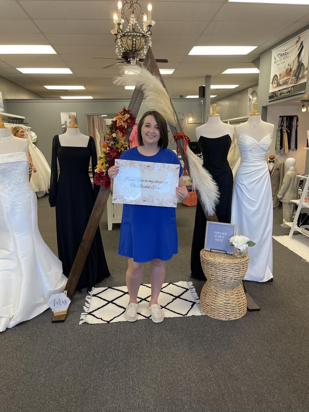 Woman in a blue dress holding a sign in a bridal shop surrounded by wedding gowns