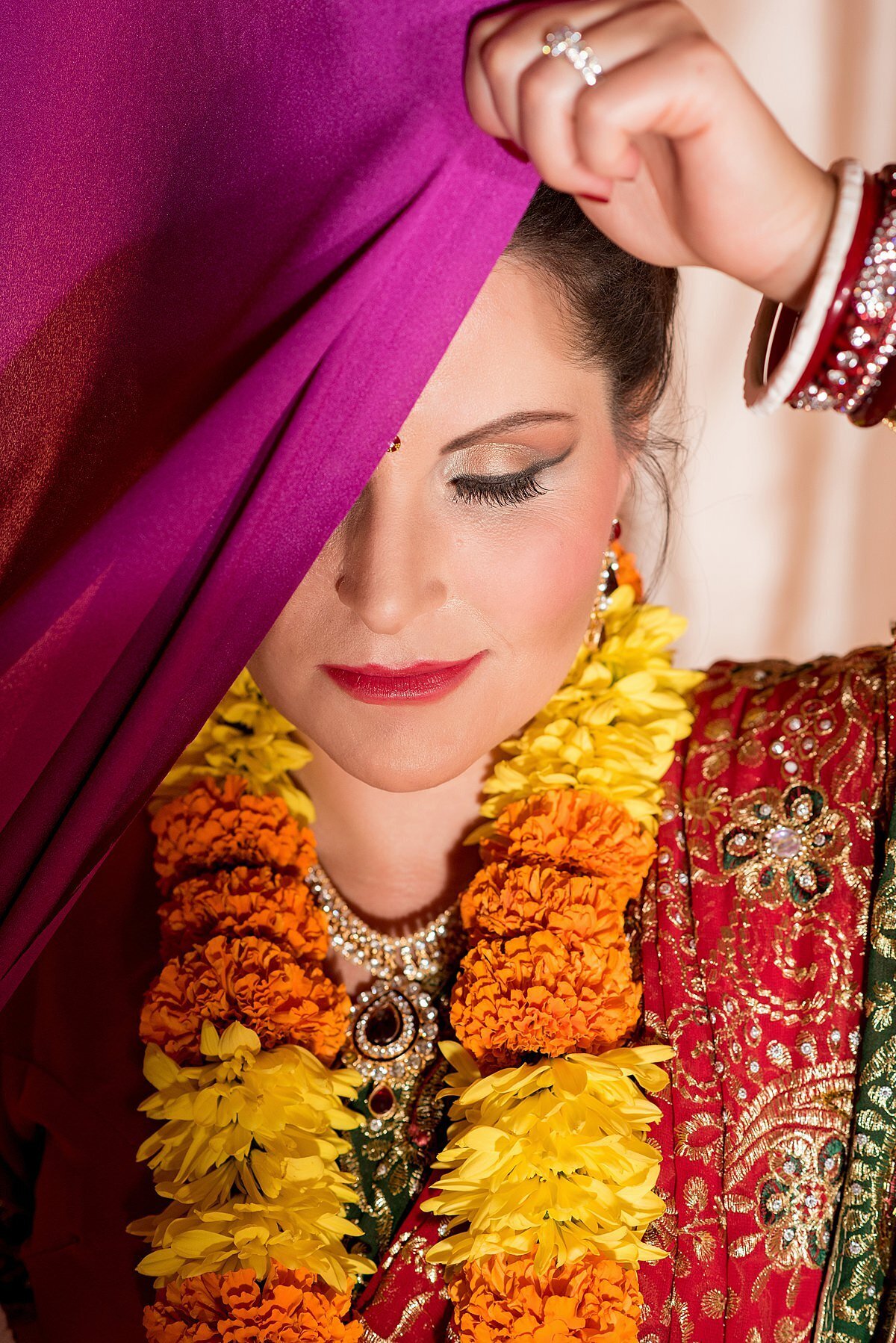 Hindu bride wearing a red and green saree with gold detail holding up a magenta dupatta. Indian bride wearing a varmala of orange marigolds and yellow mums.