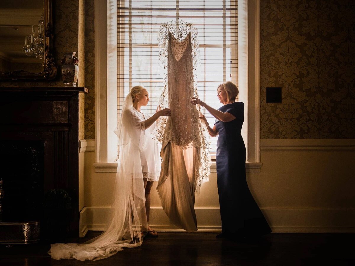 A mother and daughter share a moment of reflection as they admire a beautiful wedding gown, illuminated by the soft light of a window.