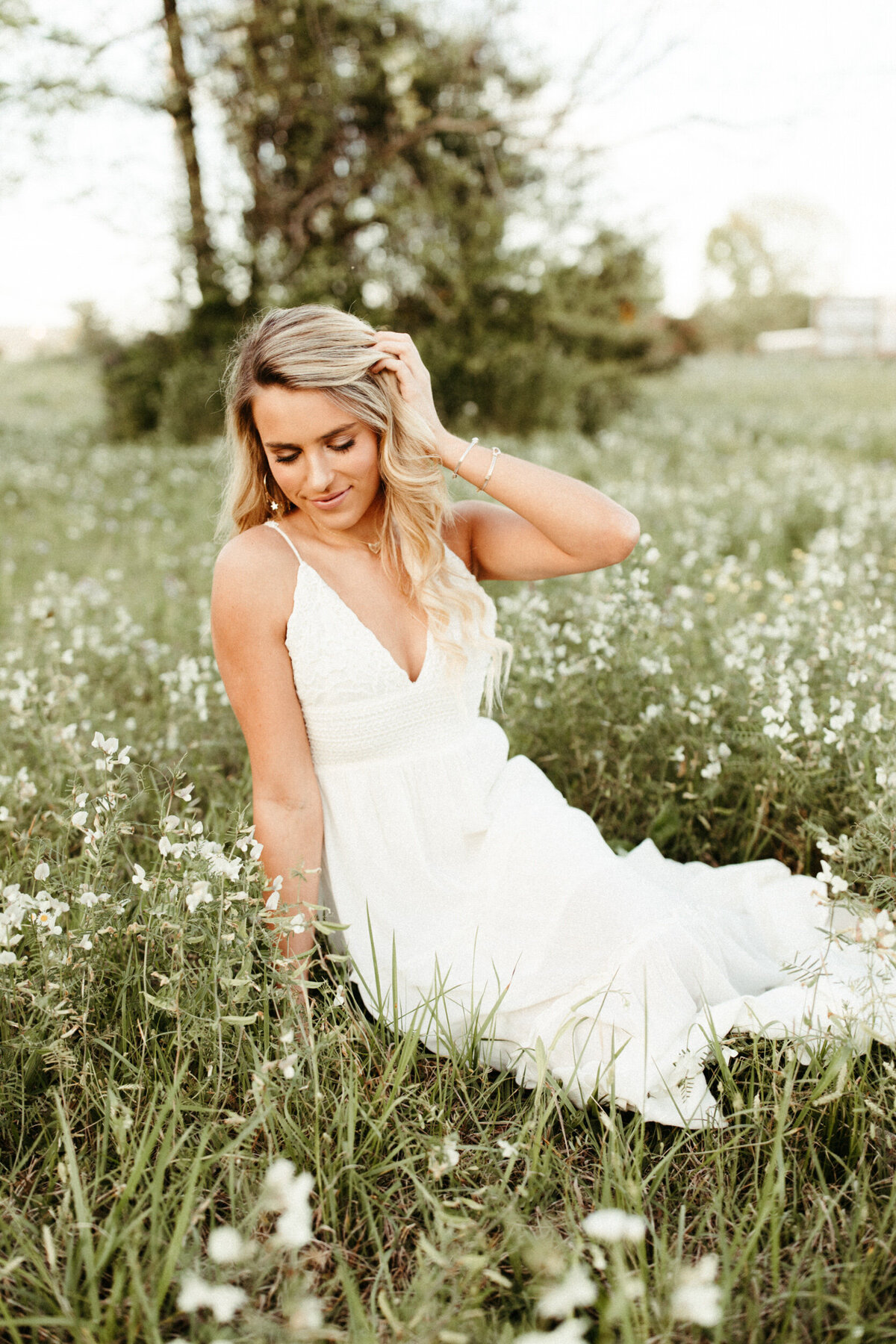 High school senior in long white boho dress sitting in a field with white wildflowers