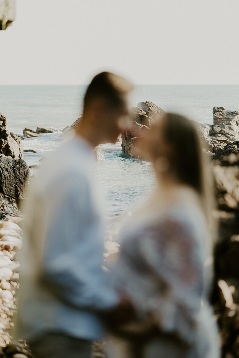 Mari et sa femme enceinte, de profil, sur le point de s'embrasser, flous. Arrière plan de mer et de rochers. Photo prise par Laura Termeau photographe professionnelle en Vendée.