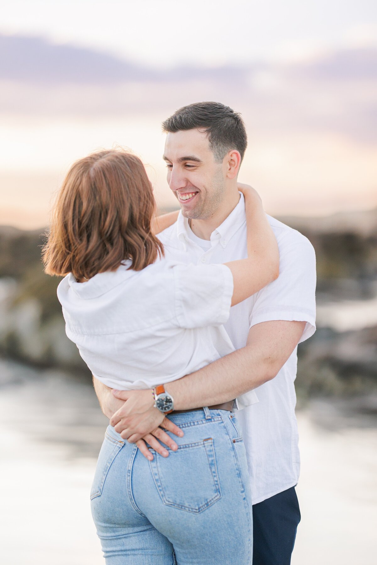 A woman with her back to the camera hugging a man and smiling during Wingaersheek Beach sunset pictures.