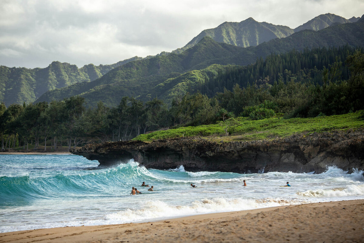 Ocean waves crash on a sandy beach with several people swimming near the shore. Dense green vegetation and tall mountains form the lush backdrop under a cloudy sky, offering a perfect setting for personalized travel planning to craft luxury vacations.