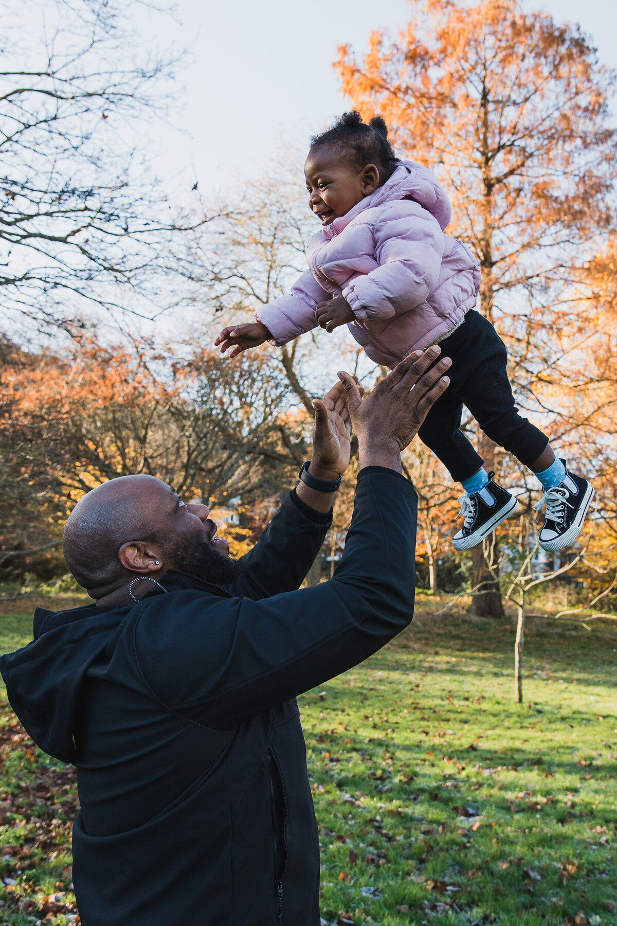 A joyful moment as a man tosses a smiling toddler in the air in a park, surrounded by autumn trees. the child wears a pink jacket and blue jeans.