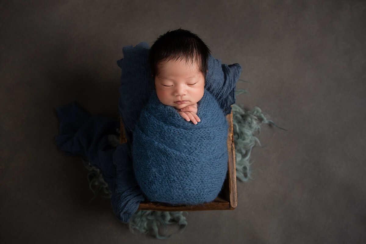 Newborn baby boy sleeping wrapped in a dark blue wrap  in a small wooden box on a grey background.