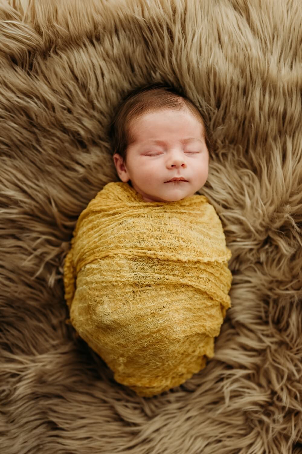 newborn sleeping on a tan fur rug wrapped in a similar colored swaddle. babys feet are sticking out.