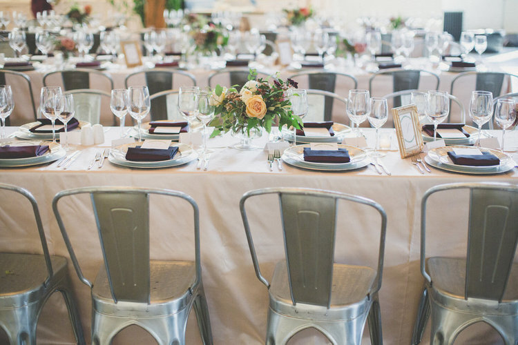 Long harvest tables with peach linens , burgundy napkins, colourful flowers and industrial dining chairs line the former art gallery called Parisian Laundry in Montreal Quebec for a wedding.
