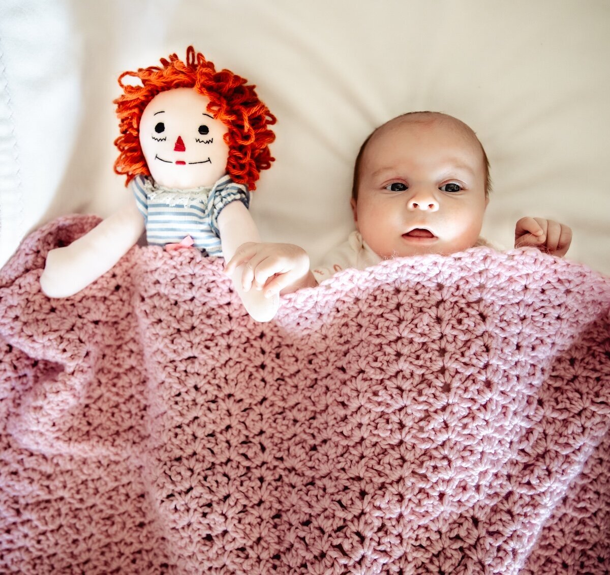A newborn baby tucked in under a pink knitted blanket next to a stuffed doll