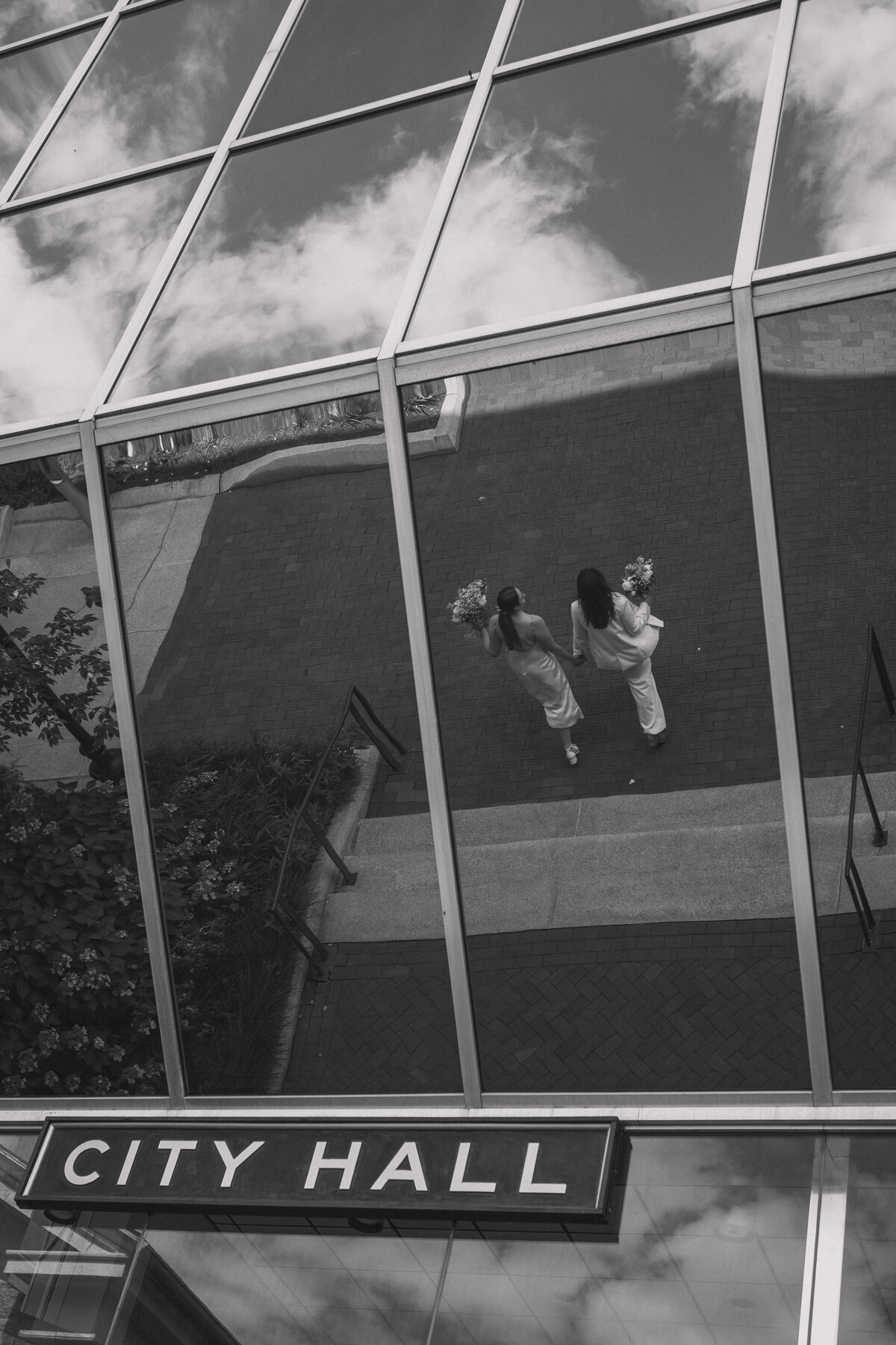 A black and white photograph capturing the reflection of a newlywed couple holding hands and walking away from City Hall. The couple, dressed elegantly, are seen from above through the reflective glass windows of the building. The sky and clouds are also reflected in the windows, adding a serene and symbolic touch to the moment. This image beautifully combines architectural elements with a candid, documentary-style wedding shot.