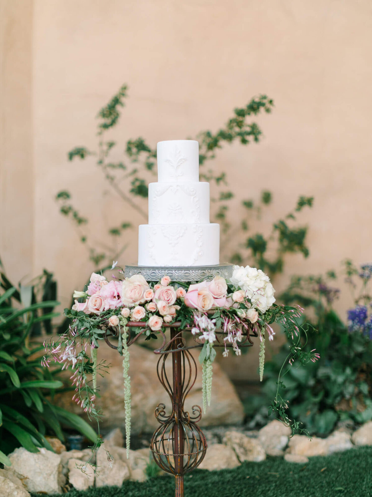 A three-tier white wedding cake with intricate patterns is displayed on a wrought iron stand.
