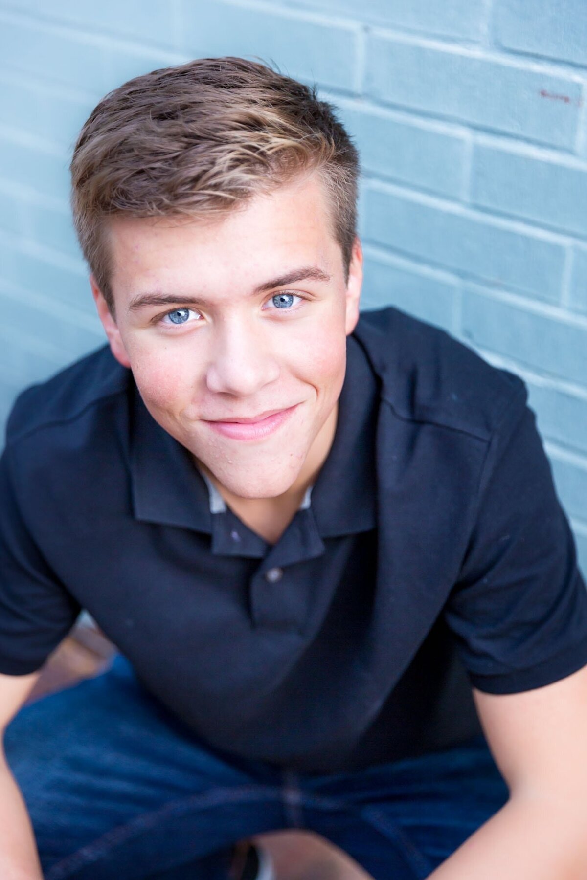 Senior portrait of a boy in a black shirt next to a blue brick wall.