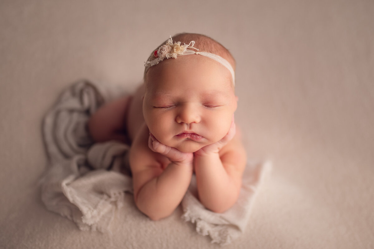 Newborn baby girl poses in our Waukesha studio, while asleep. She lays on her belly with her chin in her hands wearing a floral headband.