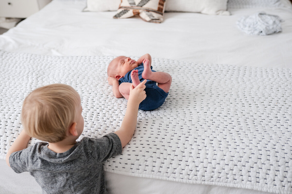 A close-up of a toddler pointing at the feet of a newborn baby lying on a bed.