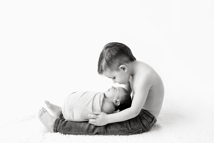 A toddler boy sits shirtless on a studio floor with his sleeping newborn baby sibling in his lap during a  NJ newborn photography session