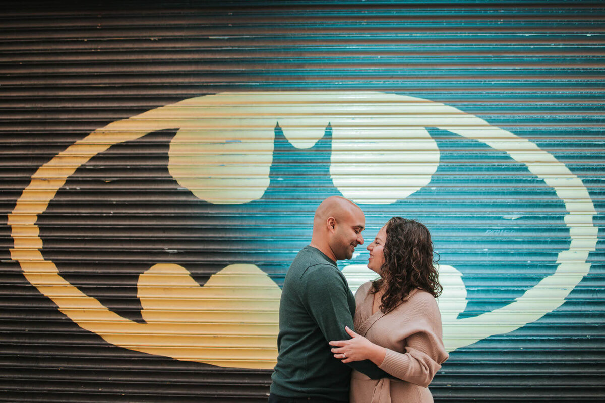 A couple embraces in front of a mural featuring a large yellow Batman logo on a metal shutter.