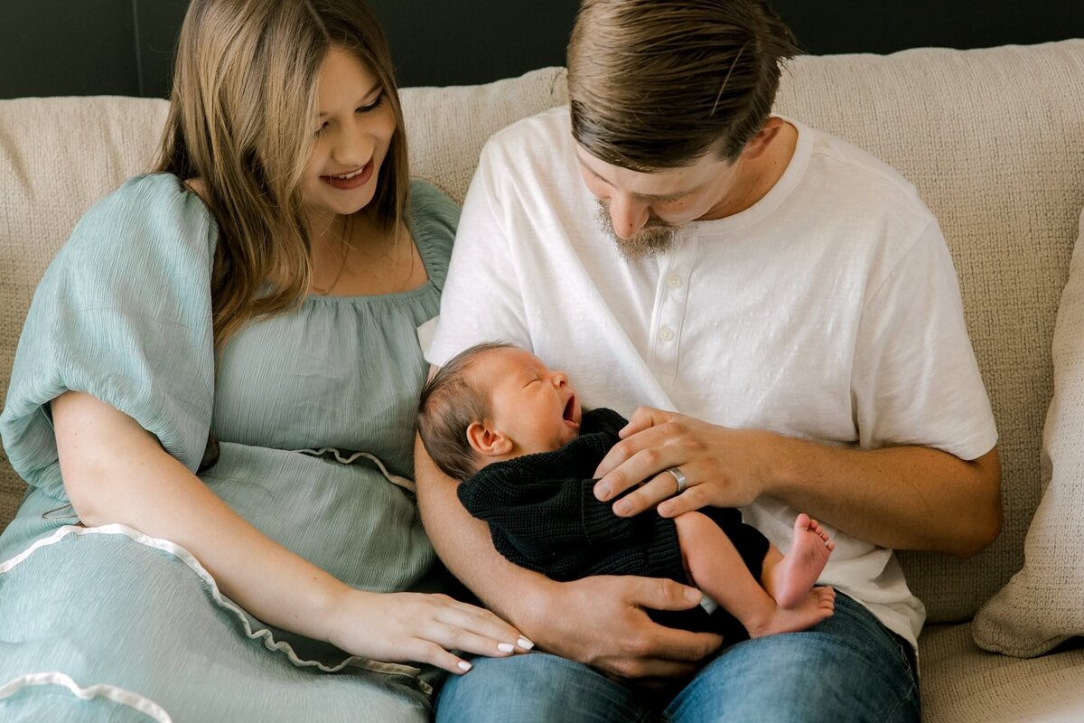 Young couple sitting on a cream-colored sectional couch, lovingly cradling their newborn baby in a cozy, natural light setting. Mary Wiebe Photography