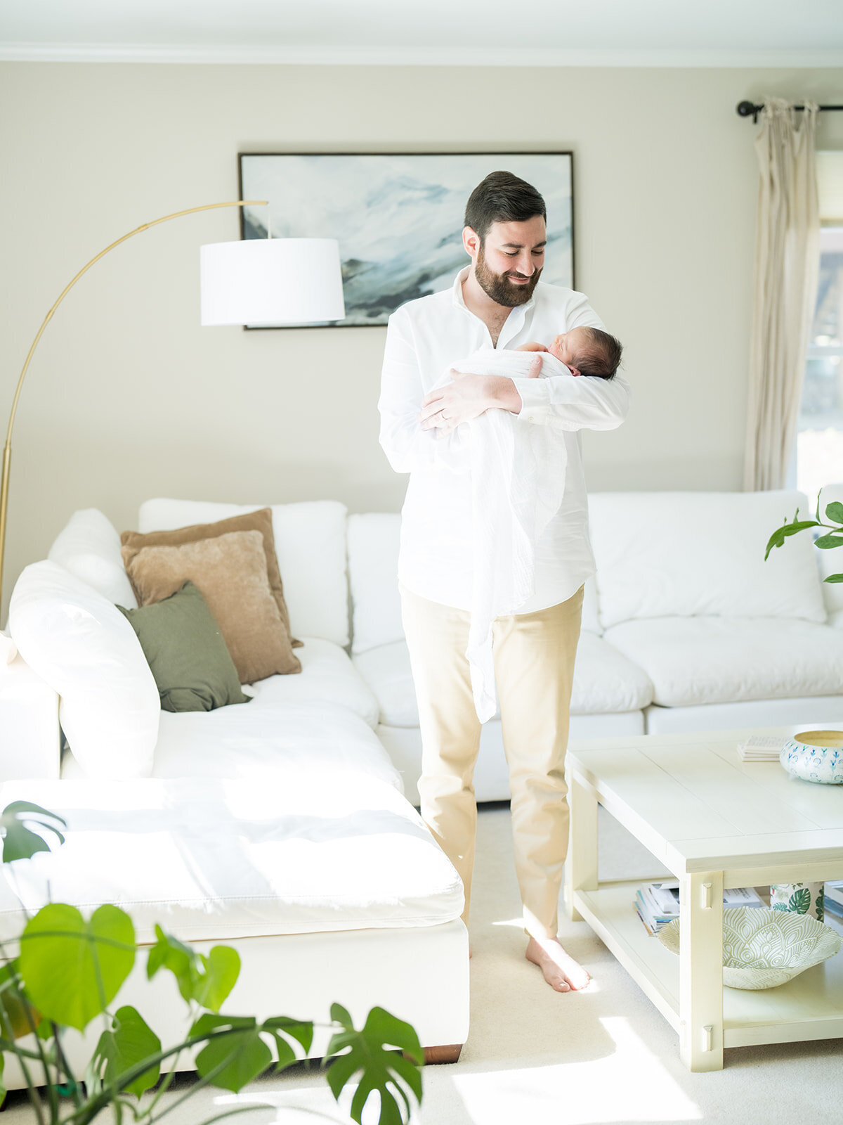 Brown-haired and bearded father stands in his white living room holding his newborn baby.
