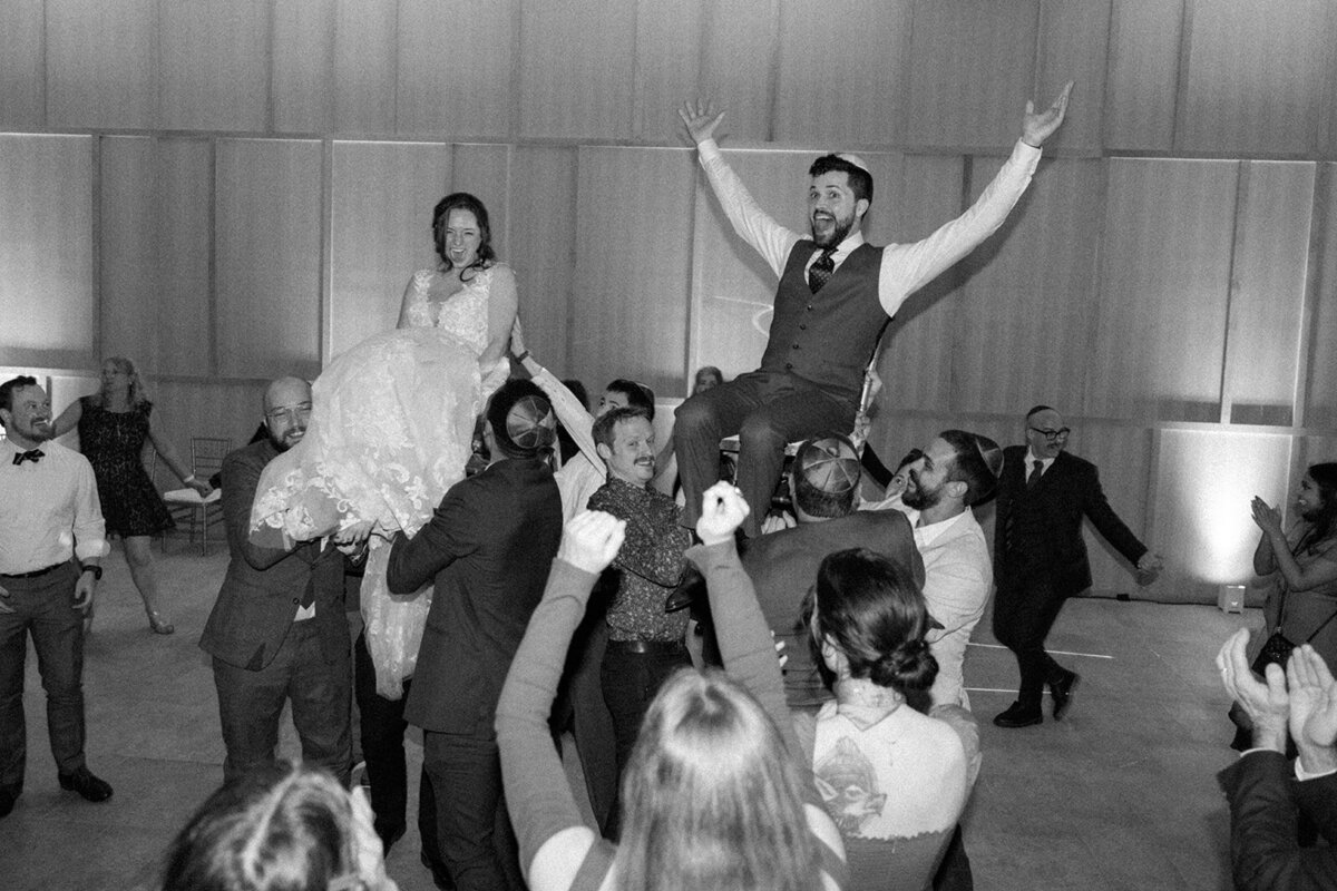 Jewish couple happily celebrates at their wedding reception while being lifted in the air on their chairs.
