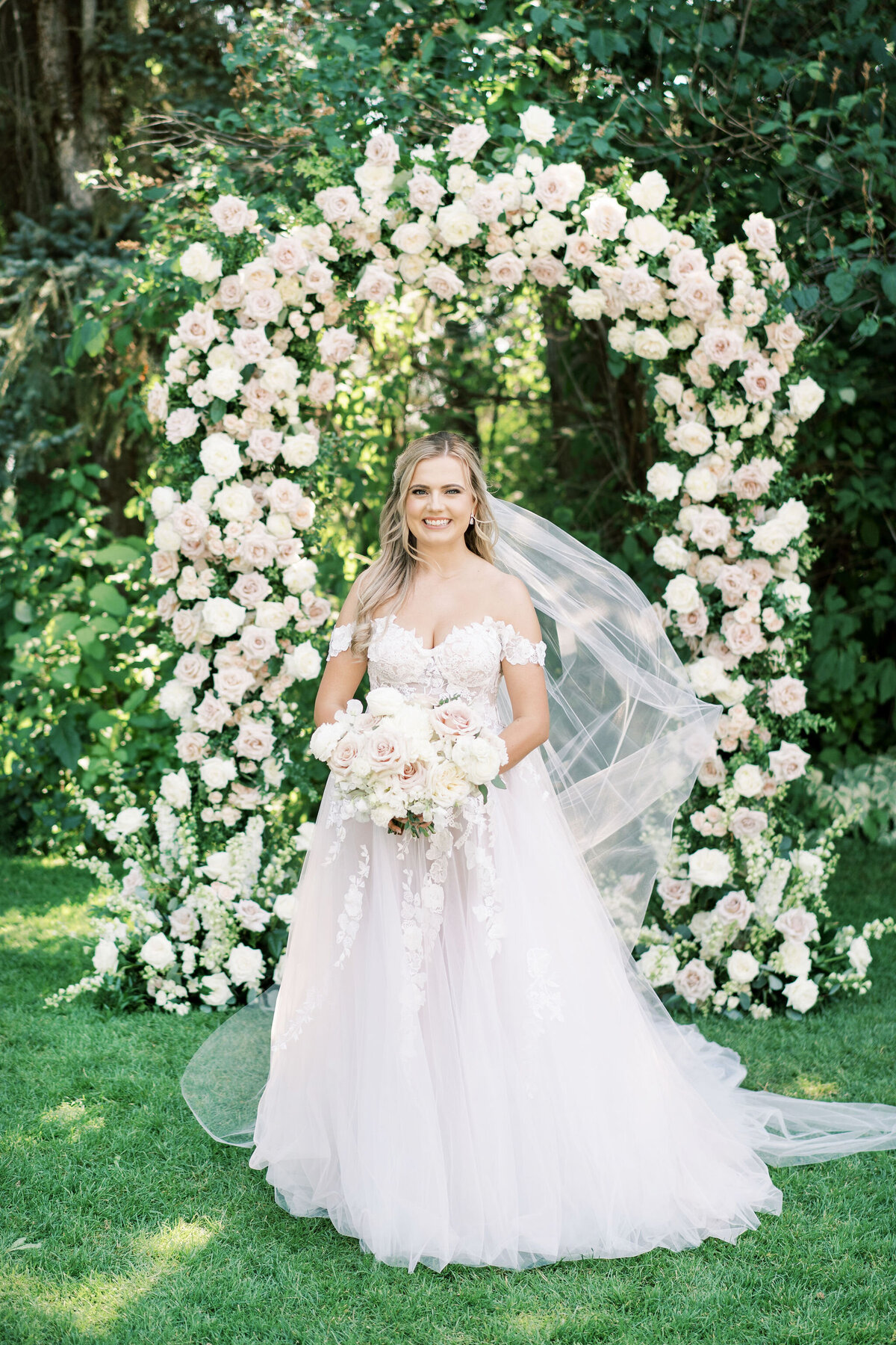 A bride in a white gown stands smiling in front of a floral arch, holding a bouquet, with the breathtaking greenery of Banff, Alberta in the background—a picture-perfect scene for a destination wedding in Canada.