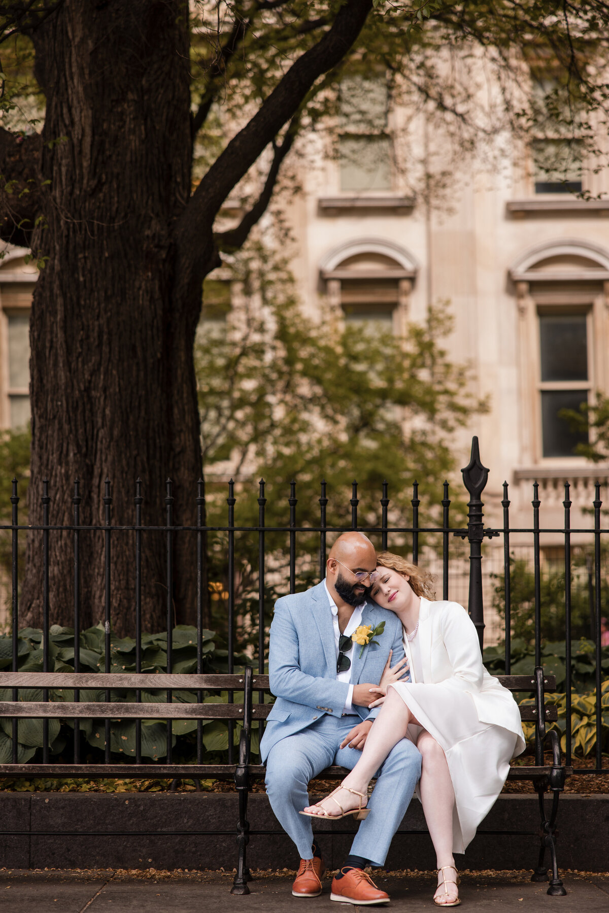 Couple seated on a bench in City Hall Park after their NYC elopement, with one partner resting their head on the other's shoulder; soft greenery and historic architecture in the background reflect the peaceful energy of a downtown Manhattan ceremony.