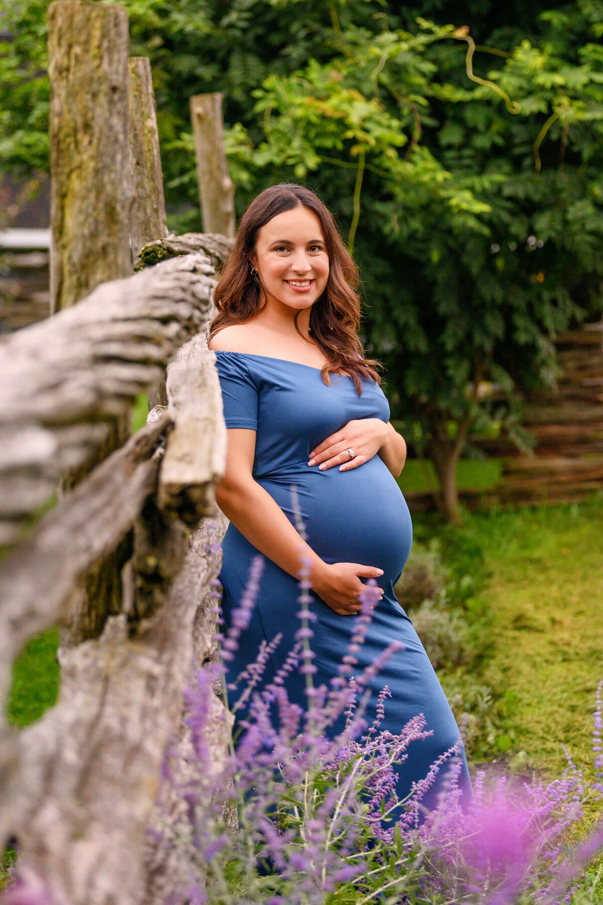 pregnant woman wearing a blue maternity dress leaning against a wood fence for her maternity photos in Burlington, ON