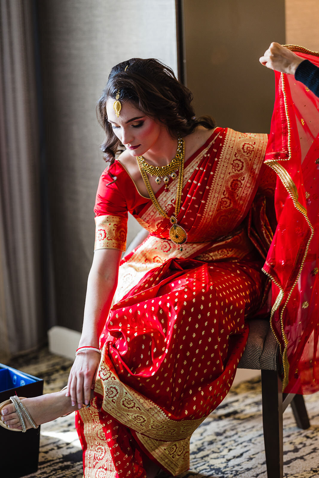 A woman dressed in a red and gold Indian saree sitting pensively with her bridal jewelry