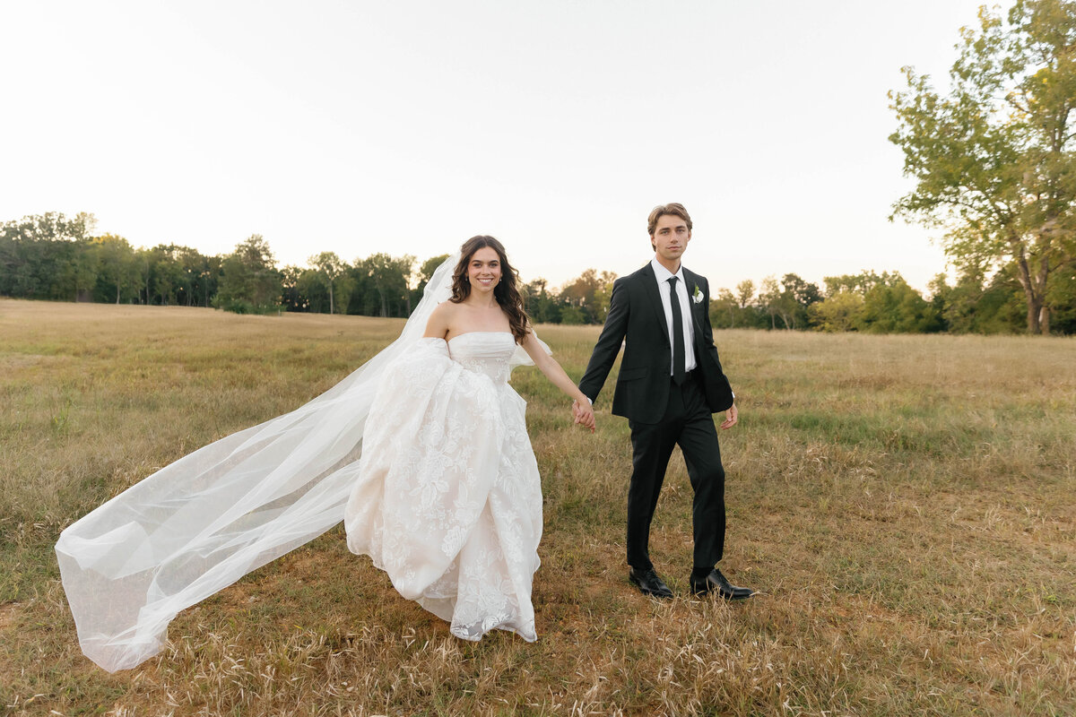 Bride and groom walking hand-in-hand through an open field at sunset, the bride’s long cathedral veil flowing behind her. She carries a romantic pastel bouquet of white and blush blooms, including roses and calla lilies. A soft golden glow highlights the couple against the natural Arkansas landscape, perfect inspiration for modern outdoor wedding florals.