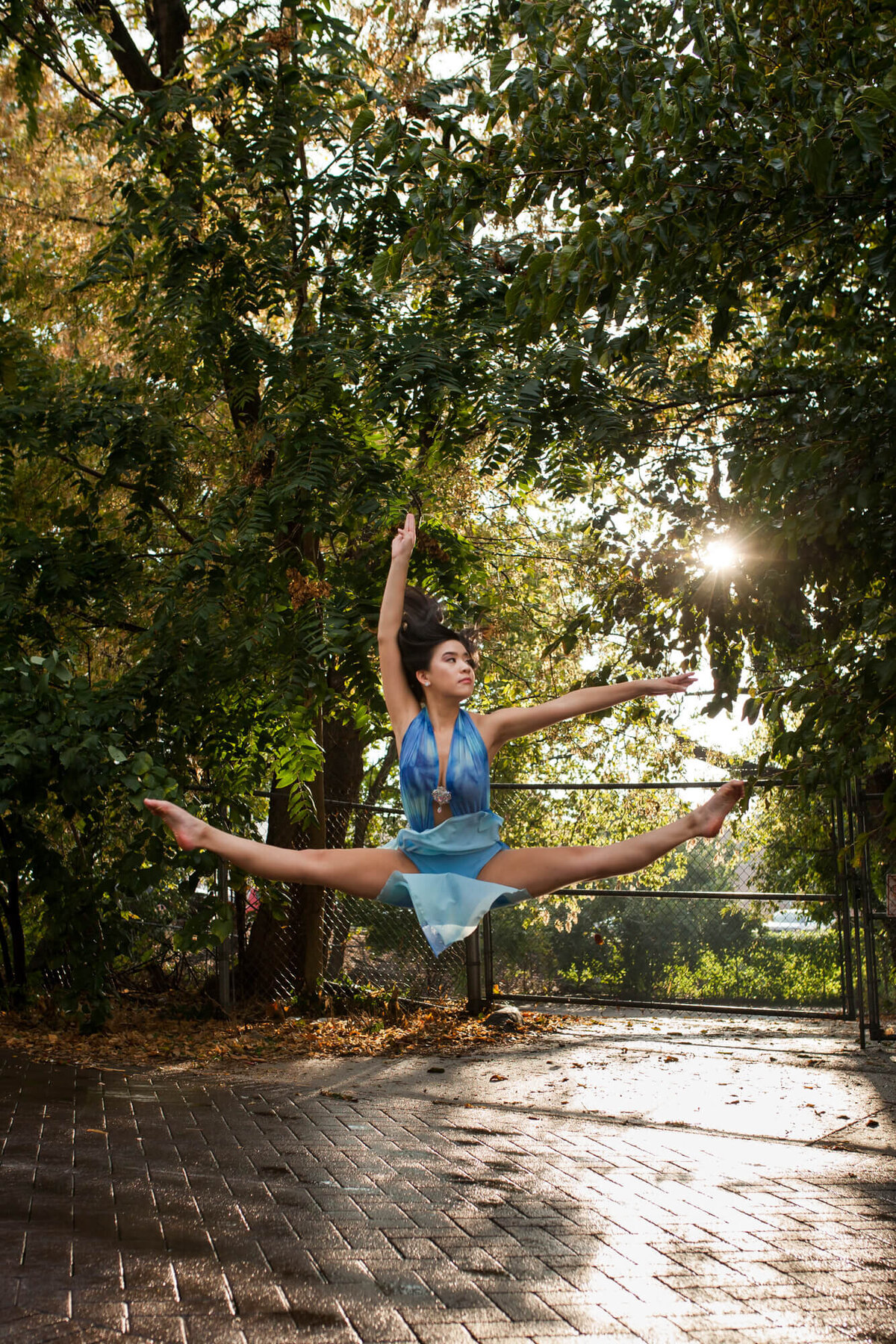 A beautiful teen dancer does a jump pose wearing a blue dance costume on a sunlit city side street. Captured by Springfield, MO senior photographer Dynae Levingston.