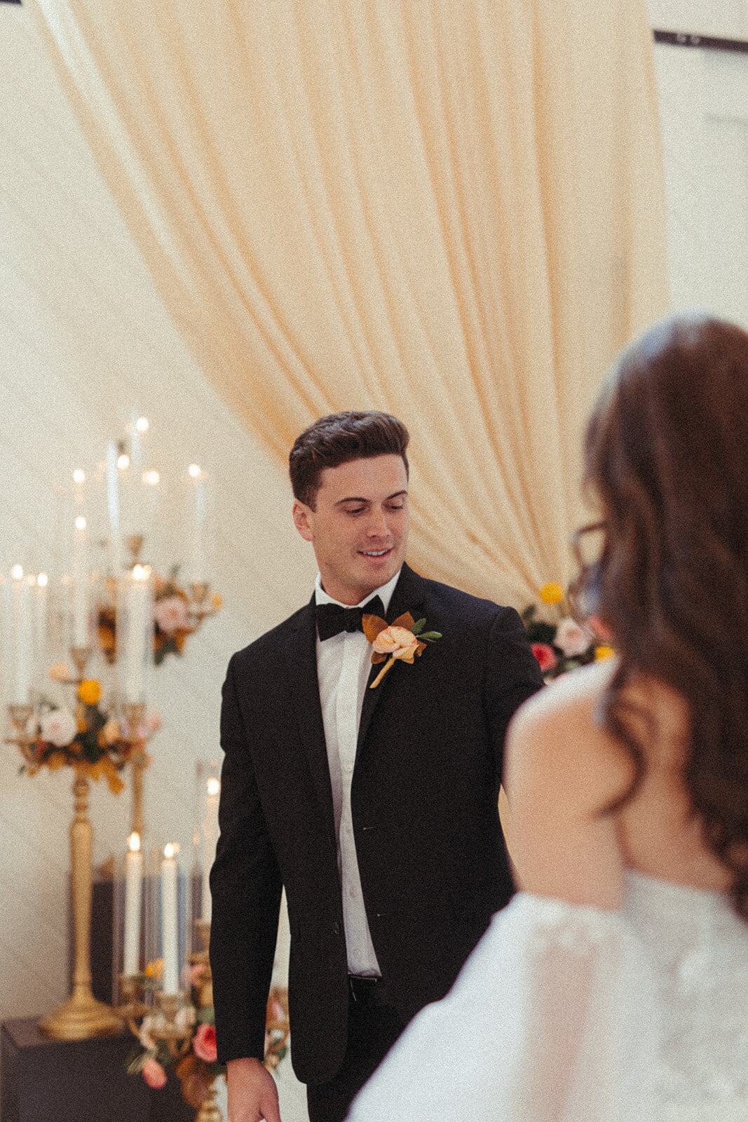 Bride and groom wearing a black tuxedo and white wedding gown standing by lit candles and peach-colored curtains.