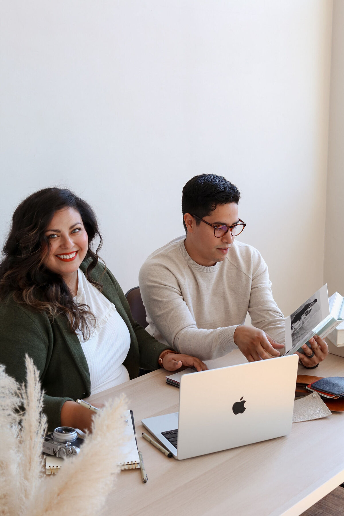 Smiling woman with lip stick  looking at camera wearing green sweater and white blouse sitting next to  man wearing light tan sweater wearing glasses looking at wedding album that he is holding- Romero Album Design