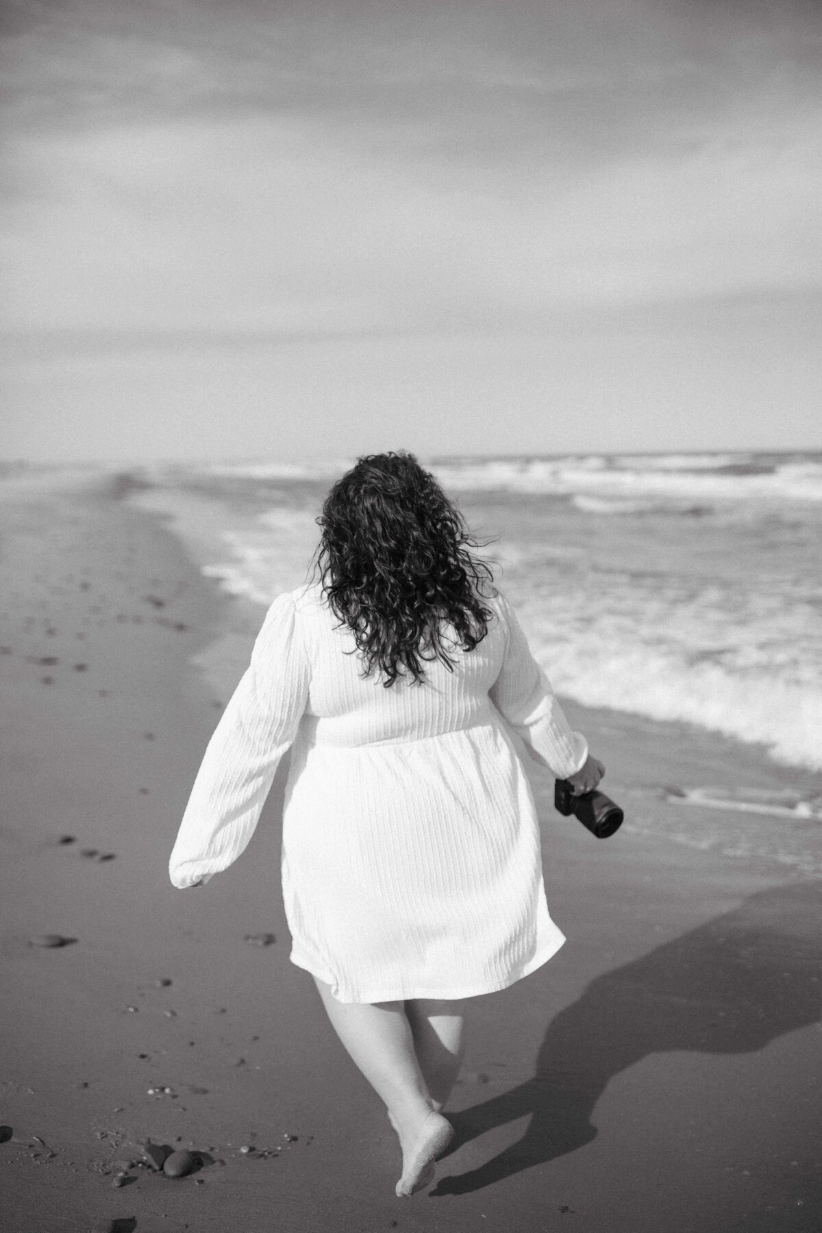 A back view shot of Valerie Doucette, a wedding, engagement, elopement, couples photographer as she walks with her camera in hand by the beach