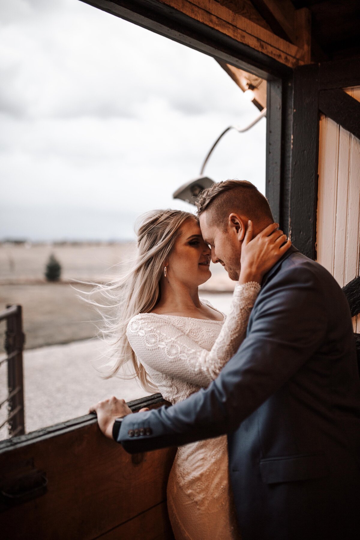 bride and groom inside of a barn together for a fall wedding with bride leaning against an open window as her hair blows in the wind while her groom leaning into her