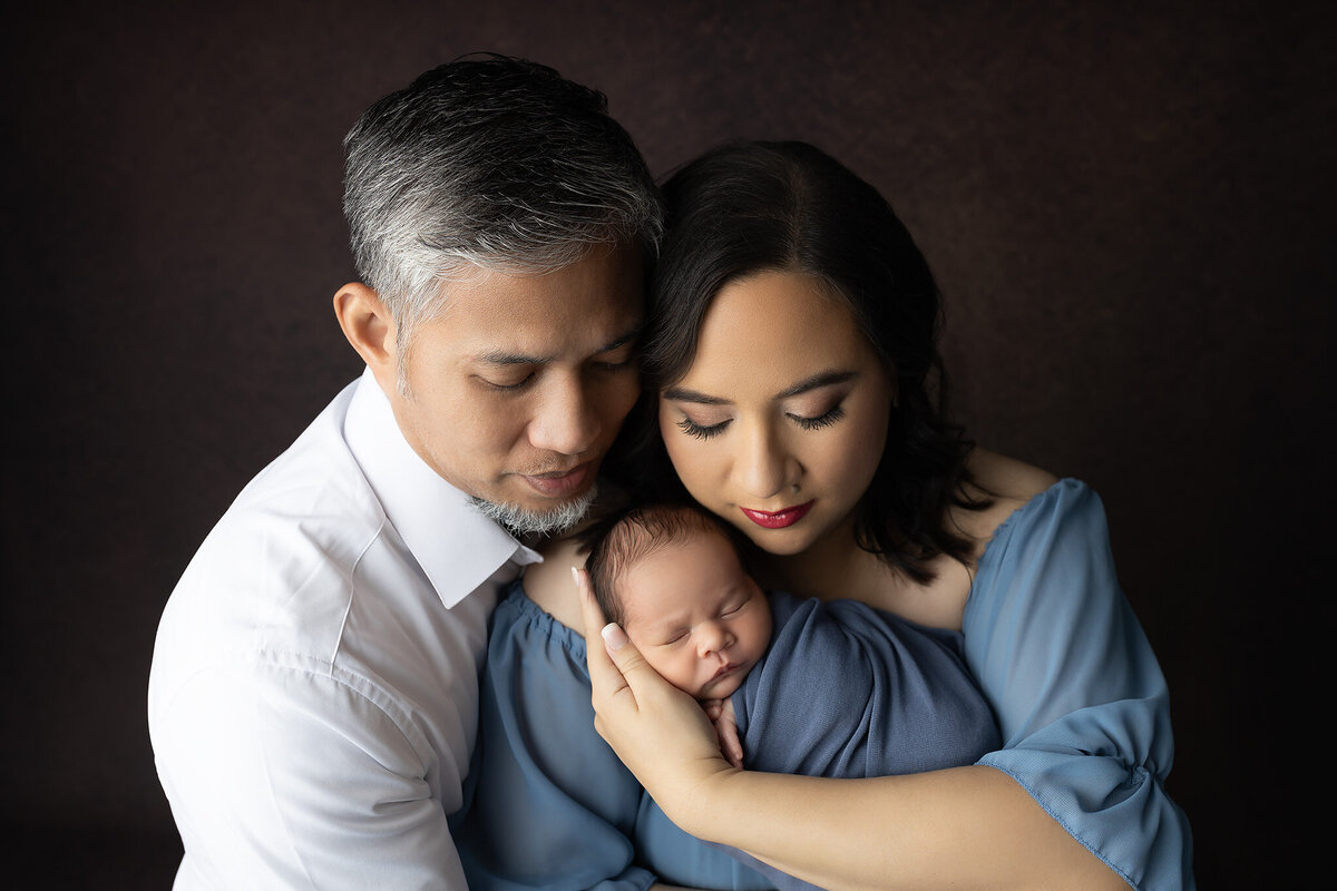 Mother and father cradle their newborn with eyes closed, embracing the calm and love of their first family portrait together.
