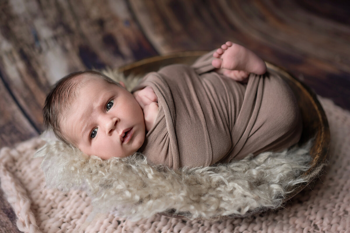 A newborn baby lays in a wooden bowl in a brown swaddle