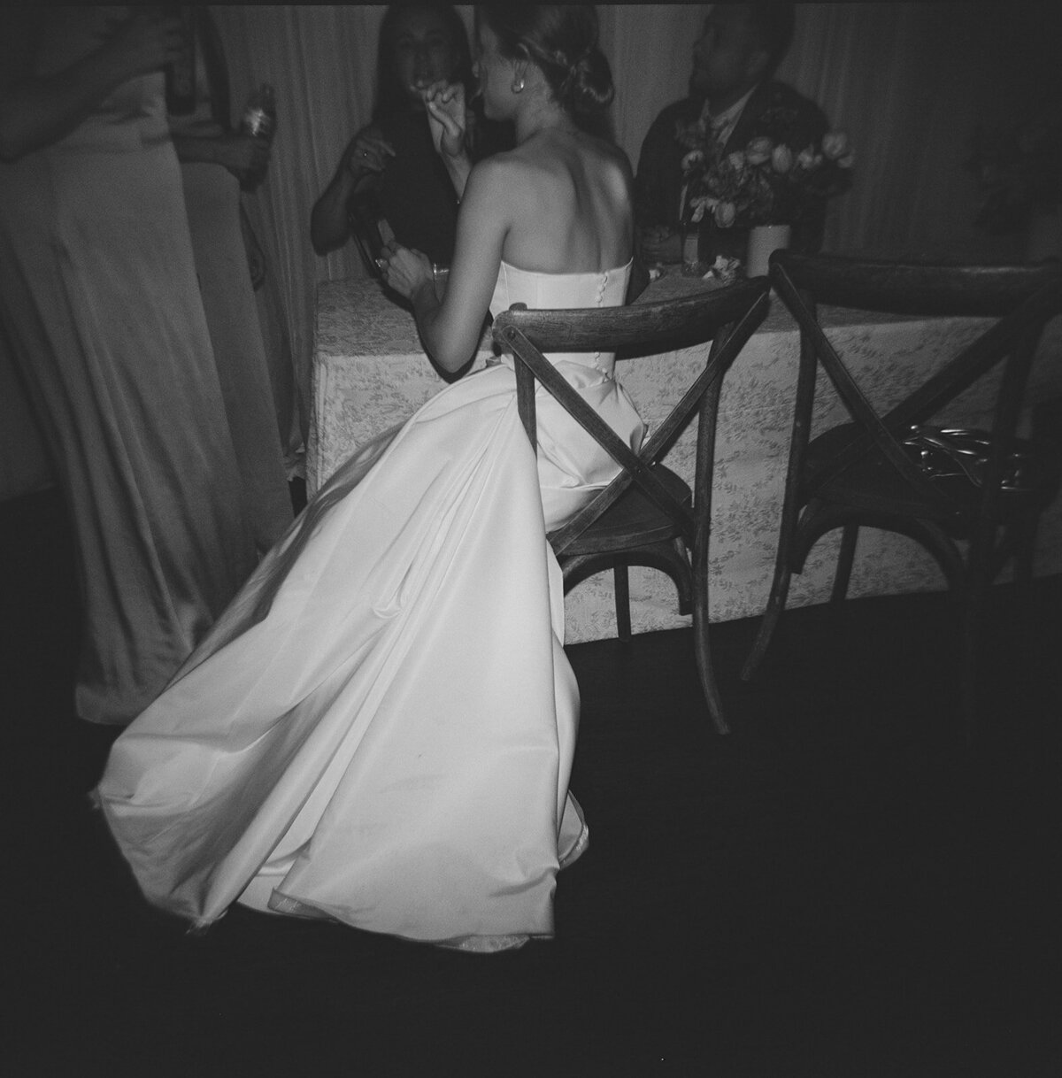 Black and white photo of the bride in a strapless ballgown sitting at a reception table, captured from behind with her train elegantly draped on the floor.