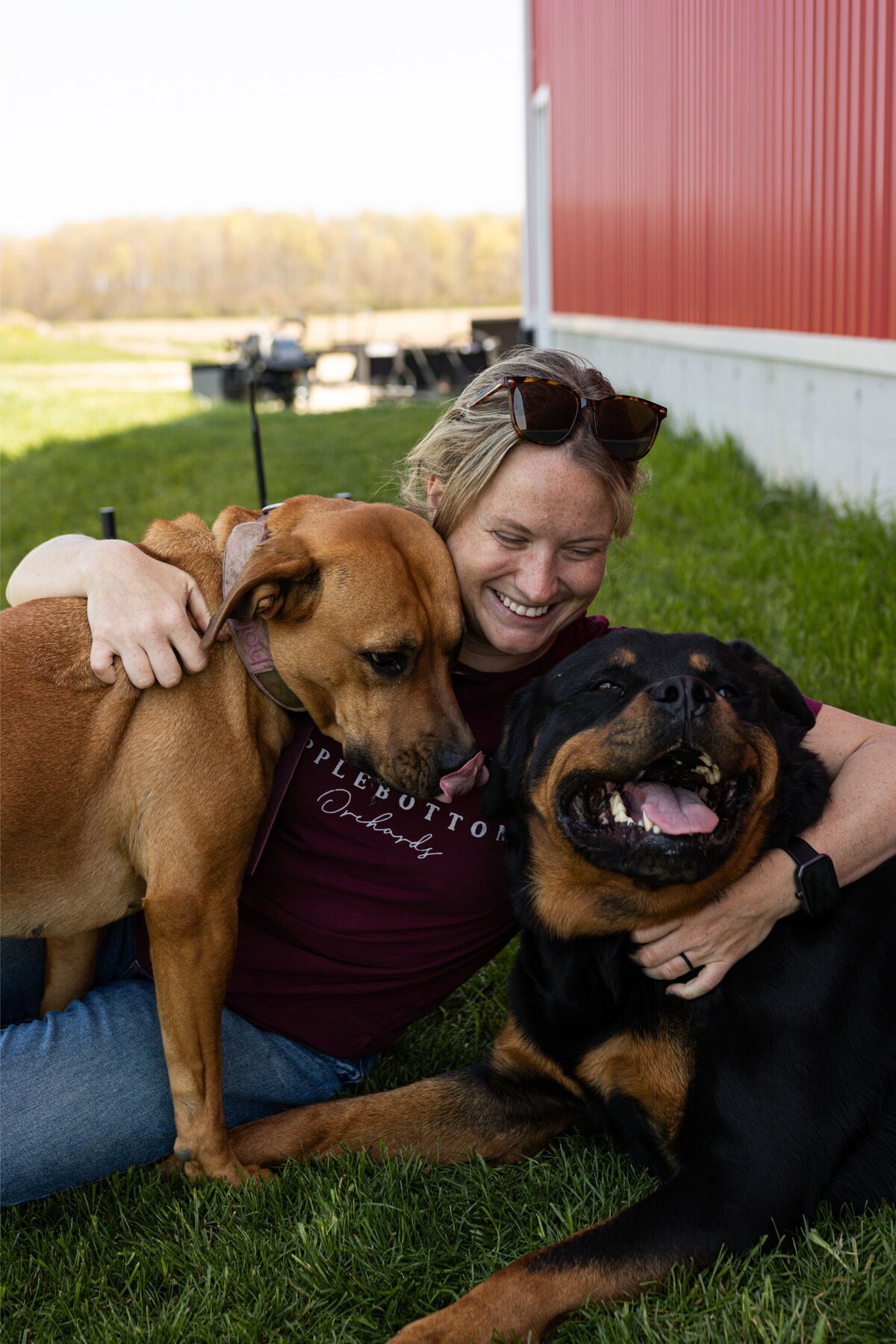 Chelsea, smiling, cuddling next to her two dogs on the ground.