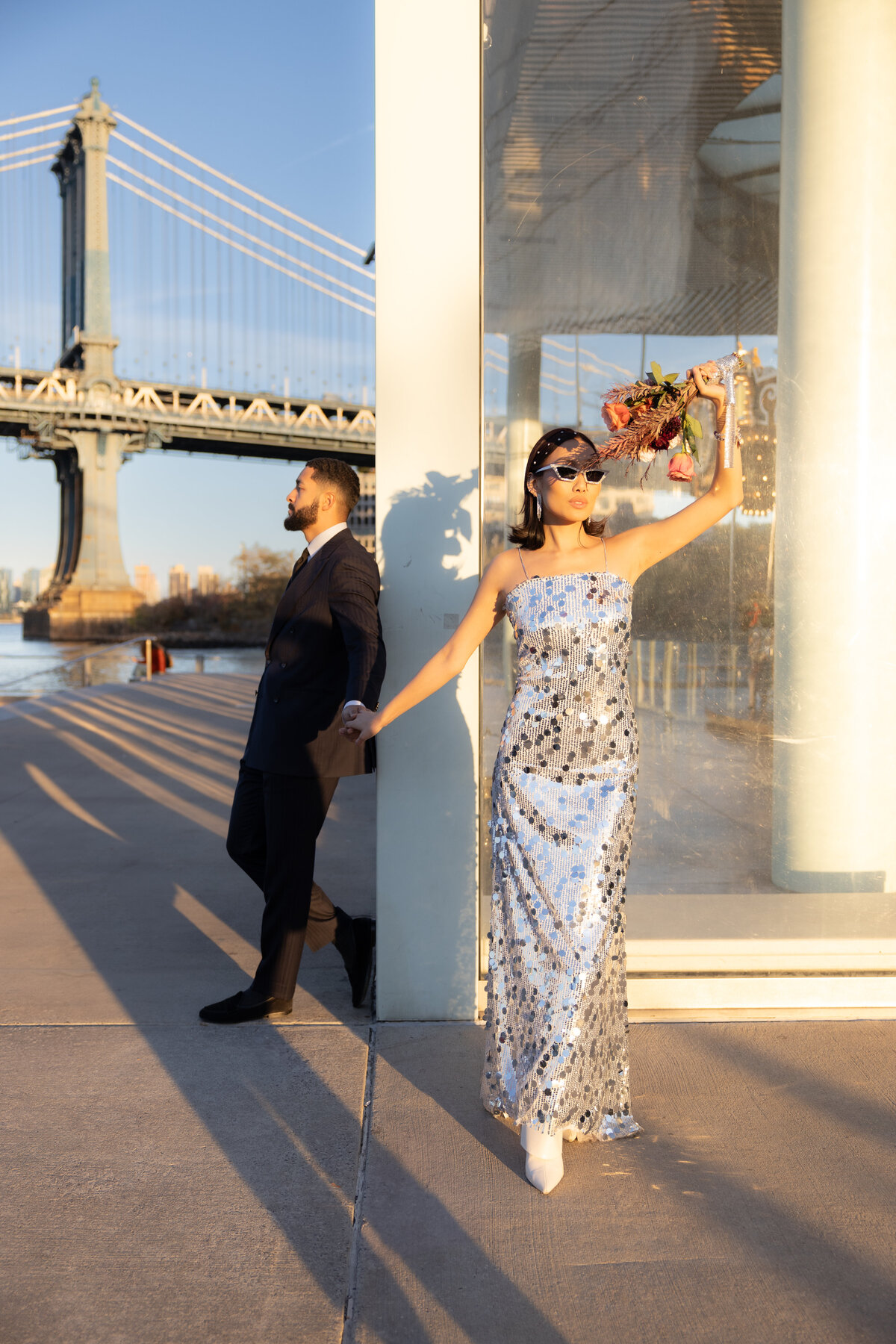 The couple poses together at the base of the Brooklyn Bridge, surrounded by urban textures and history.