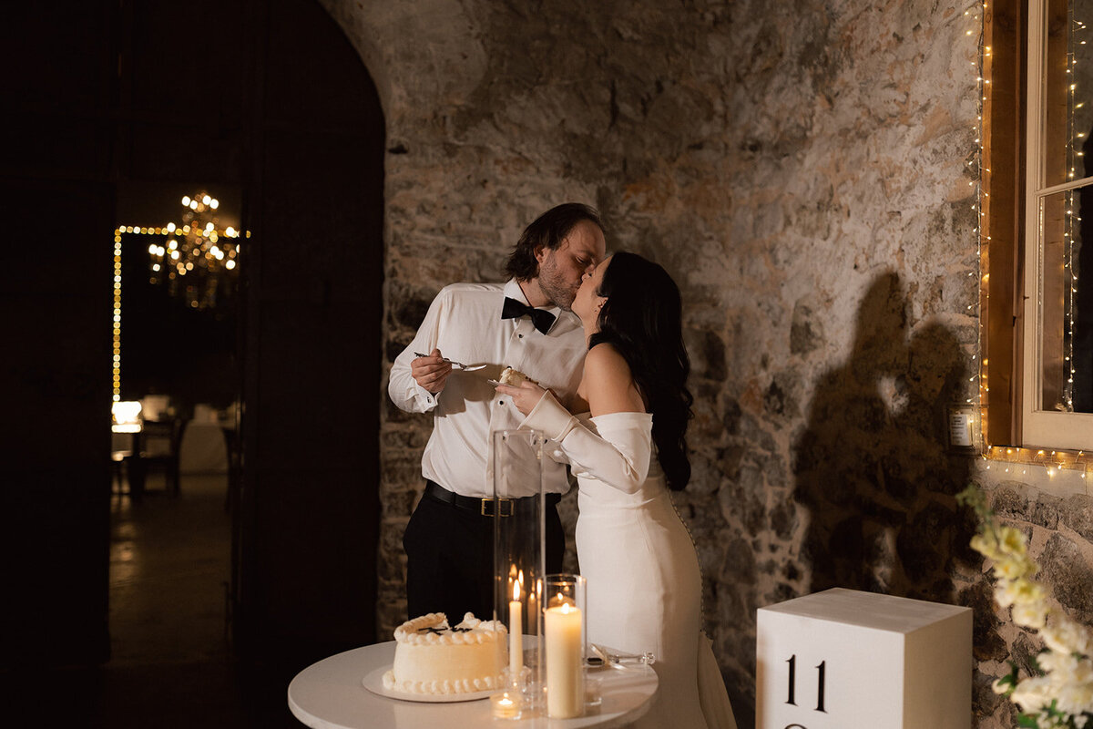A newlywed couple shares a kiss after cutting their wedding cake in a candlelit stone-walled reception. A candid and emotional wedding moment emphasizing authenticity and intimacy.