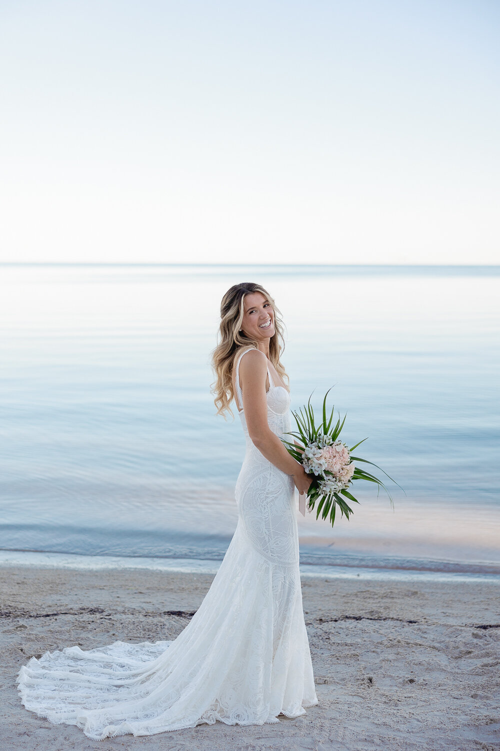 Bride Laura smiling in a stunning lace wedding gown, holding her bouquet on the beach at Cheeca Lodge Resort during her Miami destination wedding. Photographed by Claudia Amalia Wedding Photography.