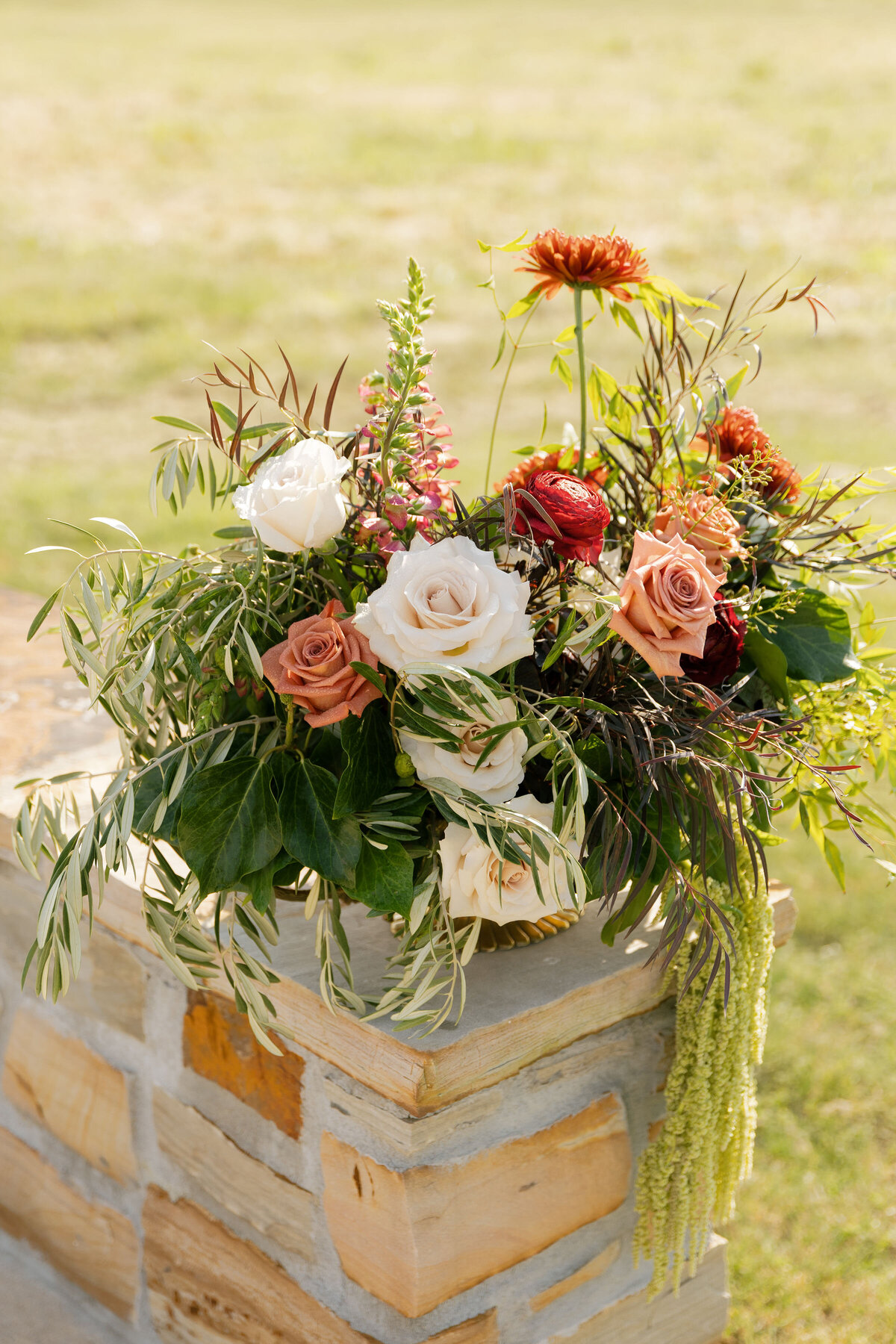 Elegant outdoor wedding arrangement with white and peach roses, autumn mums, olive branches, and cascading greenery styled on a stone ledge overlooking an open field.