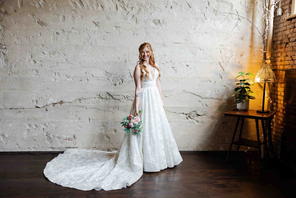 bride holding wedding bouquet down at side in front of white textured wall smiling