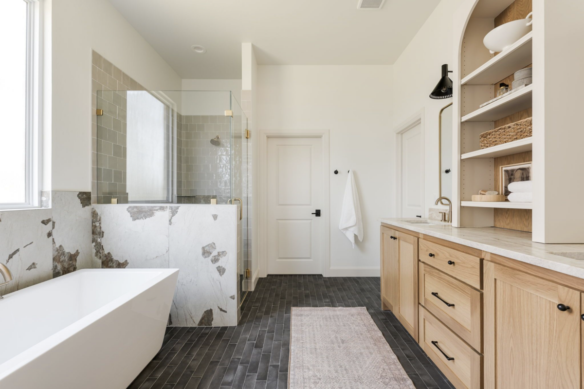 Light-filled bathroom with a freestanding tub, marble-clad tub with glass enclosured shower and brass accents, natural wood vanity with built-in shelving, and dark matte floor tile.