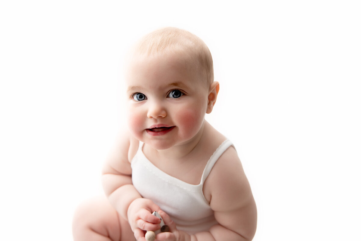 London studio high key portrait of a smiling six month old baby girl wearing a white romper.